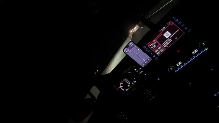 This is a first-person perspective video taken inside a car at night. The driver's hands are visible on the steering wheel, and the car's dashboard displays are illuminated. A smartphone is mounted on the dashboard, showing a navigation app. The car's headlights illuminate a road ahead, with other vehicles visible in the distance. The video appears to be a recording of a drive at night.