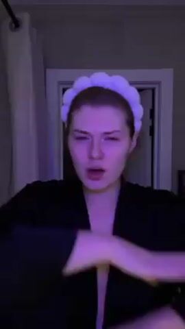 The video shows a person in a bathroom. The person is wearing a black robe and a light-purple puffy headband. They are making various hand gestures and mouthing words, presumably singing or rapping along to music. The bathroom has a white door slightly ajar and a shower curtain on the left. The lighting in the room is predominantly purple.