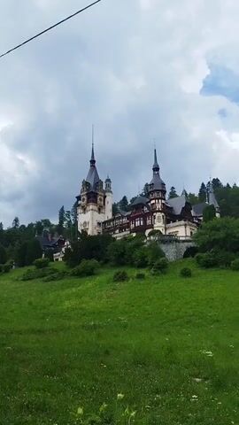 The video shows a castle on a hill with a cloudy sky. The castle is a large building with multiple towers and spires. It is surrounded by trees and a lush green lawn. The camera pans slowly from left to right, giving a panoramic view of the castle and its surroundings. The overall scene is picturesque and serene.