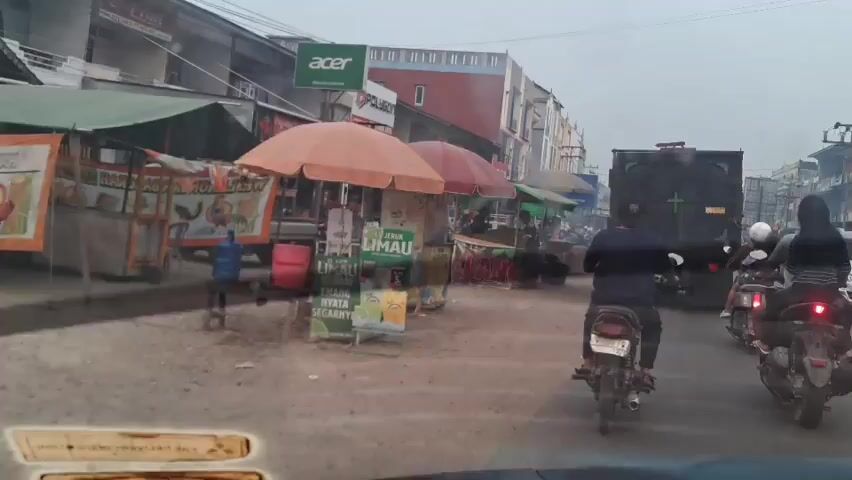 The video shows a street scene with several food stalls on the left side of the road. The stalls have umbrellas providing shade. One stall has a sign with the word "LIMAU" and images of fruits. Another stall has a sign with the word "LANG" in large letters and images of what appear to be potatoes. People are seen standing and working at the stalls. On the right side of the road, there are vehicles including a dark colored truck and several motorcycles. Some people are riding the motorcycles, one of whom is wearing a black hijab. The road appears to be unpaved and dusty. Buildings line the street in the background, some with signs that are partially visible. The sky is overcast. The overall impression is a bustling street market.