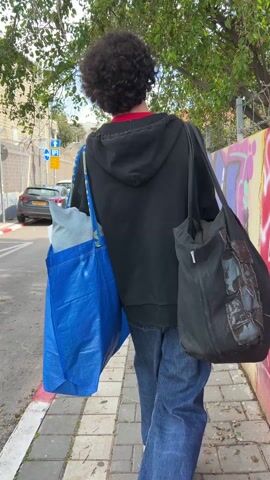 A person with curly hair is walking down a sidewalk. They are wearing a black hoodie, red shirt, and blue jeans, and carrying a blue Ikea bag on one shoulder and a black tote bag on the other. The sidewalk is paved and next to a street with parked cars. There's a graffiti-covered wall and green trees in the background.