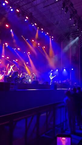 A concert is in progress. A band is playing on a stage, illuminated by stage lights. The stage features drums, guitars, and multiple performers. The lighting is dynamic, with vibrant blue and purple hues dominating, interspersed with yellow and red spotlights. A crowd is visible in the foreground, watching the performance. The overall environment suggests an outdoor or large indoor venue.
