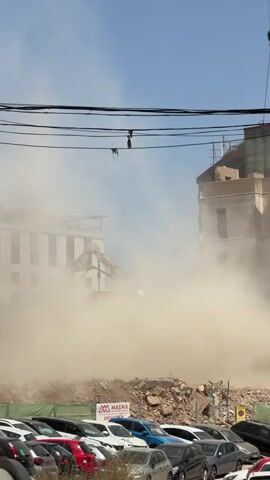 The video shows a demolition site on a sunny day with a clear blue sky. In the foreground, a large amount of dust hangs in the air, obscuring much of the scene. Below the dust cloud, a row of parked cars is visible, mostly white and some other colors. Behind the cars, a green construction fence and a pile of rubble can be seen. In the middle ground, a large excavator with a yellow boom is partially visible through the dust, actively engaged in demolition. Further back and to the right, a tall building is in the process of being demolished; its upper floors are intact, but the lower sections are crumbling, with debris falling and contributing to the dust. A crane with a hook is also visible above this building. To the left, another building, lighter in color and partially obscured by dust, appears to be under construction or renovation. Overhead, black electrical wires crisscross the frame. The overall impression is one of ongoing construction or deconstruction, with a significant amount of dust generated by the heavy machinery.