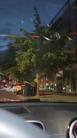 This is a first-person perspective video from inside a car at dusk. The camera is pointed forward through the windshield, showing a street scene. There are trees with green leaves lining the sidewalk. To the right of the trees, there is a building with large windows. On the sidewalk, there are several large black trash bins with yellow lids. Several cars are visible on the street, some of which are moving and some are parked. Traffic lights are visible and appear to be red. The sky is dark blue with some clouds, and a sliver of the moon is visible. The interior of the car, including the dashboard and steering wheel, is partially visible in the foreground. The overall impression is of a city street at the end of the day, with cars and pedestrians moving about.