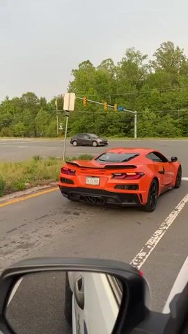 The video shows the rear of a bright orange Corvette stopped at an intersection. The car has a Virginia license plate that reads "DA GAINZ." The Corvette has a prominent rear spoiler and four exhaust pipes. The intersection has traffic lights and a "One Way" sign visible. Trees and foliage are visible in the background. The video is taken from inside another car, with the side mirror visible in the lower left corner of the frame.