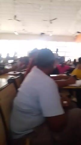 The video shows a classroom full of students seated at desks. The room is brightly lit by natural light coming through a series of windows along one wall. The ceiling is white with multiple ceiling fans. There is a colorful mural on one wall of the classroom. The students are facing forward, and appear to be engaged in studying or taking a test.
