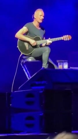 A man with gray hair and a black t-shirt is playing a black acoustic guitar while sitting on a stool. He is wearing black pants and appears to be on a stage with blue lighting. There are speakers in the foreground.