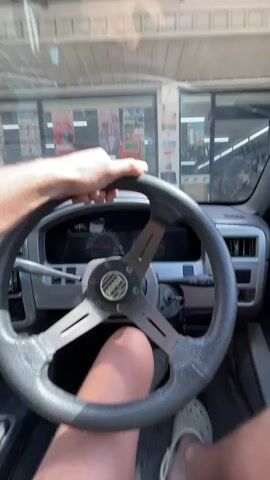 The video begins by showing the interior of a car from the driver's perspective. The driver's hands are on the steering wheel, which is a sporty aftermarket model. The dashboard and instrument cluster are visible. The camera then pans to the right, showing the car's interior door panel and the side mirror. The view shifts to outside the car, revealing a convenience store with glass doors and windows. The store appears to be a 7-Eleven, indicated by the prominent logo. The car is parked in front of the store, with yellow parking lines visible on the ground. The driver's leg and foot, wearing a light-colored Croc shoe, are also visible in the frame. The video ends as the camera continues to look towards the store entrance.