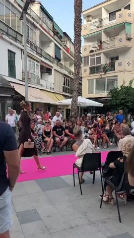 A fashion show is taking place on a street. A model in a brown dress walks down a pink runway. Spectators are seated on chairs on either side of the runway, facing inwards. Buildings with balconies and palm trees line the street. The sky is clear and the sun is setting.