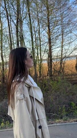 The video shows a young woman walking along a path. She has long brown hair and is wearing sunglasses, a white hoodie, and a beige trench coat. She is walking away from the camera and looking to her right. She turns to look briefly at the camera. The background consists of trees with bare branches and reeds. There is water visible in the distance and the sky is clear and blue.