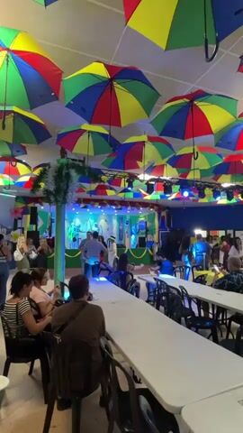 The video captures an indoor event space, likely a restaurant or hall, filled with people and decorated festively. The most prominent feature is the ceiling, which is densely covered with numerous open umbrellas in bright primary colors: red, yellow, blue, and green.

At one end of the room is a stage where a live band is performing. Musical instruments, including drums, a guitar, and a keyboard, are visible. The stage is adorned with green and yellow decorations, possibly banners, and lit with stage lights that occasionally change color. Several people are gathered in front of the stage, dancing and enjoying the music.

Throughout the rest of the room, white rectangular tables with black chairs are arranged for seating. Many individuals are seated at these tables, observing the performance, socializing, eating, and drinking. Their attire appears casual. The lighting in the venue is ambient, complemented by the stage lights, suggesting an evening event. The overall atmosphere is lively and celebratory. No discernible text is visible within the video frames.