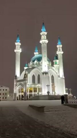 The video begins with a static, centered, eye-level shot of a white mosque with blue domes and minarets at night. The mosque is illuminated with white and gold lights. There is snow on the ground and a few people are visible near the mosque. The camera then quickly pans down to the ground, which is covered in snow, before quickly tilting up to show a man standing on the snowy ground. He is wearing a black jacket and a black beanie and he is waving. The mosque is in the background.