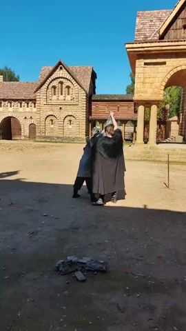 Two individuals are engaged in a mock fight outdoors in what appears to be a historical reenactment village. One person, dressed in a grey tunic and trousers, is being held by the other, who wears a long black cape with a fur collar and dark boots. The person in the cape has their right arm raised, seemingly in a striking or grappling motion. The background consists of rustic stone and wood buildings with thatched roofs under a clear blue sky. The ground is dusty dirt. The interaction appears to be staged, with no signs of genuine distress.