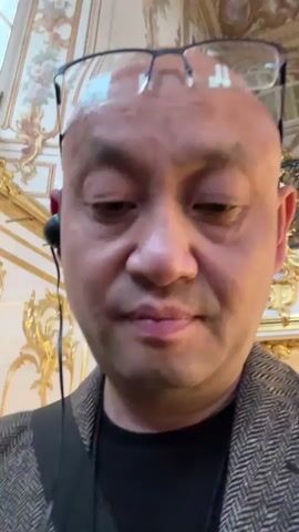 This is a close-up, first-person perspective video, likely filmed on a smartphone. The primary subject is a person's head and upper body, with a focus on their face. The individual is bald and wearing glasses perched on top of their head, along with earbuds. They are dressed in a dark t-shirt and a textured, herringbone-patterned jacket.

The background suggests an opulent interior, with ornate gold detailing visible on what appears to be a staircase railing and architectural elements. This decorative style, combined with the location context of Peterhof, Russia, points towards a grand palace or museum setting, possibly during daytime given the visible light. The person's expression is neutral, perhaps thoughtful or observing, and their gaze is directed downwards and slightly to the side. The activity seems to be a casual recording while exploring the location. No visible text is present in the video.