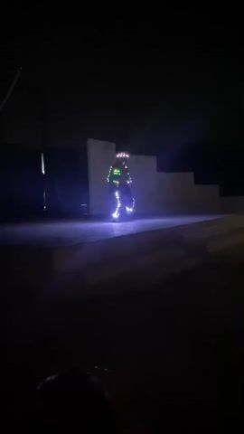 A person in a dark costume with glowing LED lights stands on a dimly lit stage at night. The stage is concrete and has a set of steps in the background. A large metal hoop is visible on the left side of the stage. The overall scene is dark.