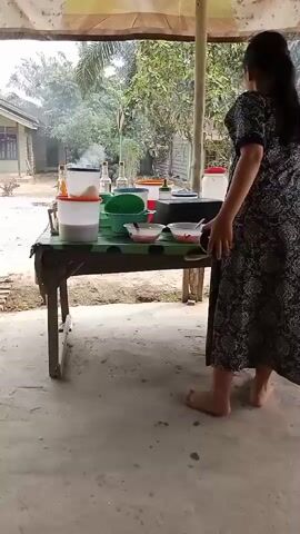 A woman wearing a dark-colored dress with a floral pattern stands facing away from the camera, looking towards a table. The table is covered with a green cloth and holds various items including containers, bowls, and a small stove with smoke rising from it. To the right of the table, a red machine with a silver component is visible. The scene appears to be outdoors, with trees and a building in the background. The woman is barefoot.