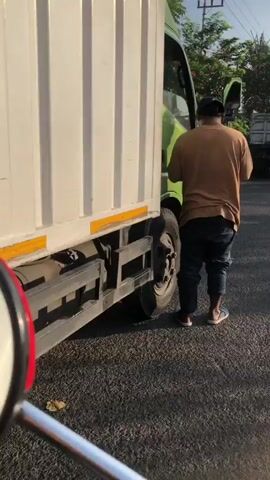 The video shows a man standing next to a white box truck. He is wearing a brown t-shirt, blue jeans, and sandals. He is facing away from the camera. The truck is parked on a paved road. The sun is shining, and there are trees in the background. The image is taken from a low angle, possibly from a motorcycle.