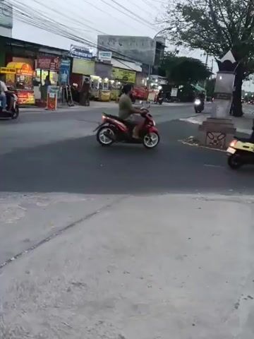 The video captures a street scene with several motorcycles in motion. On the left, a motorcycle with a rider wearing a white helmet and a dark jacket is turning left. Further down the street, another motorcycle is moving away from the camera. In the center of the frame, a motorcycle with a rider wearing a light-colored shirt is heading towards the right. To the right of that, a pink scooter with two occupants, one in a pink shirt, is traveling towards the viewer. Behind the pink scooter, a motorcycle is moving away. Another motorcycle is visible in the far background, on the right side of the street, heading away from the camera. The environment appears to be a commercial area with shops and food stalls lining the left side of the road. There are utility wires overhead, and a tree with dark foliage stands on the right side of the road near a monument or decorative structure. The road surface is asphalt, and there is a concrete sidewalk or pedestrian area in the foreground. The lighting suggests it might be dusk or early morning.
