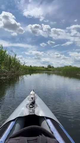 The video begins with a close-up view of the front of a gray inflatable kayak moving through a narrow river or canal. Tall, green reeds line both banks of the waterway. The sky above is blue with scattered white, puffy clouds. The water is calm and reflects the sky and surrounding vegetation. The camera is positioned low, giving a first-person perspective of someone kayaking. The kayak's bow is visible at the bottom of the frame, with a black backpack or bag behind it. The video shows a continuous forward motion through the tranquil watery environment under a partly cloudy sky.