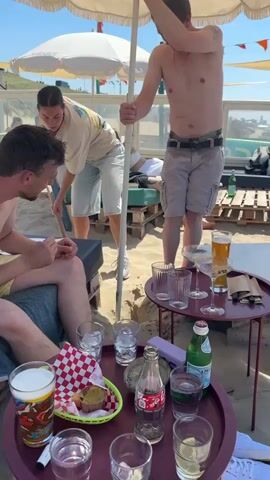 The video captures a group of people on a sunny beach. In the foreground, a person is sitting on a lounge chair. A reddish-purple tray with drinks and snacks is placed on a table in front of them. Behind the seated person, a woman is using a shovel to dig a hole in the sand near a white umbrella pole. A man stands next to her, holding the umbrella pole. In the background, other people are visible lounging on chairs and the beach. The scene includes a beach setting with sand, umbrellas, and the ocean in the distance, viewed through a glass barrier.
