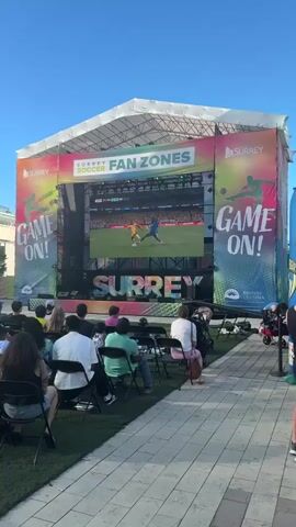 A large outdoor screen displays a soccer game with the score 0-0 in the 77th minute between ECU and CHN. Two players are on the field, one in a yellow and black uniform is on the ground, and another in a blue uniform is standing over him. Below the screen, the word SURREY is displayed in large, colorful letters. A crowd of people sits on chairs watching the game. The event appears to be a fan zone event.