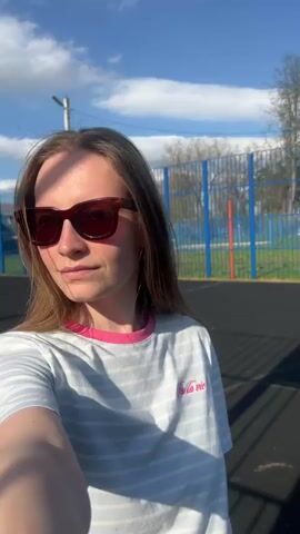 The video shows a young woman taking a selfie video outside on a sunny day. She has long brown hair and wears sunglasses, a white and blue striped t-shirt with a pink collar and a pink inscription. She is standing in what appears to be a basketball court with a blue fence in the background. The sky is blue with scattered clouds. The woman smiles at the camera and sticks her tongue out. The video is rotated at one point, then returns to the initial view.