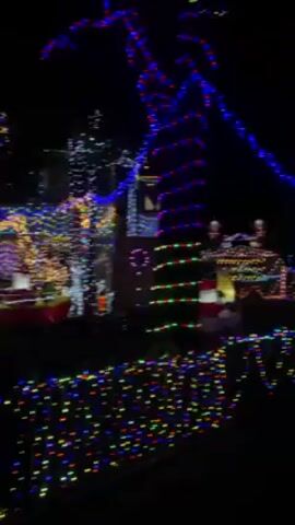 The video showcases a house adorned with vibrant Christmas lights at night. The camera pans across the decorated yard, capturing a multitude of multicolored lights outlining the house, trees, and yard fixtures. Towards the end, a person walks into the frame and briefly raises their arm towards the illuminated house. The surroundings are dark, with glimpses of parked cars and a street light. The scene conveys a festive and bright atmosphere.