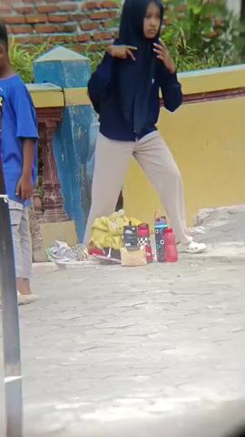 The video shows a group of children in an outdoor setting, possibly a schoolyard or playground. In the foreground, a child in a white t-shirt and grey shorts is facing away from the camera, with their right arm extended forward. To the left, another child in a blue shirt and grey pants is looking down, appearing to be walking. Behind them, a group of children are gathered, some wearing t-shirts and others with headscarves. There are also several objects on the ground, including a yellow bag, water bottles, and what appear to be shoes. The background features a yellow and blue wall and some greenery. The overall impression is of a casual gathering of children.