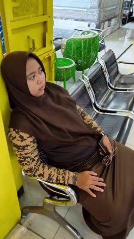 A person wearing a brown hijab and an orange shirt stands behind a counter, holding a phone and interacting with a payment terminal. In the background, another person in a purple hijab is visible near a display. The environment appears to be a retail store, possibly selling furniture, with products and signage displayed. The lighting is bright, and the overall scene is of a transaction taking place.