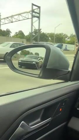 The video, taken from inside a moving vehicle, shows a black Dodge Challenger reflected in the side mirror. The Challenger is traveling in the same direction, appearing to be in the lane behind. The video provides a view of the multi-lane highway, with various other vehicles visible in the background. Trees and highway signage are also visible along the side of the road. The interior of the recording car is partly visible, including the door panel with a silver handle and red light.