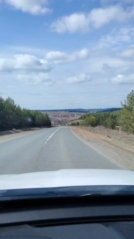 The video captures a first-person perspective from a vehicle driving on a paved road in Zabegalovo, Russia. The road extends straight ahead, sloping gently downwards towards a valley where a cluster of houses and buildings, indicative of a village or small town, is visible in the distance. The road features a white line marking the center. Tall evergreen trees, likely pines, line both sides of the road, creating a natural border to the landscape. Beyond the trees and the distant settlement, the terrain appears flat or gently rolling, stretching towards the horizon. The sky is bright blue with numerous scattered white, fluffy clouds, suggesting clear and pleasant daytime weather. No specific activity beyond driving is captured, and no people or visible text are present in the frame. The scene evokes a sense of open road travel through a rural or semi-rural landscape.