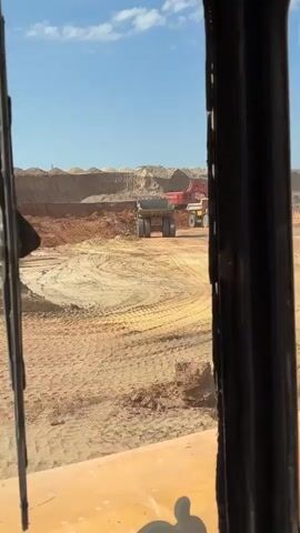 The video shows a view from inside a vehicle looking out at a construction site. There are two large trucks and a red excavator in the distance. The ground is sandy and uneven, with piles of dirt and sand. The sky is blue with some clouds. There are also some metallic structures visible in the foreground, possibly part of the vehicle the camera is in.