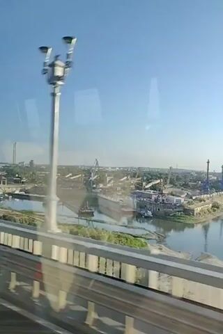 The video shows a view from a bridge overlooking a river and a port. The sky is clear and blue. On the left side of the screen, there is a dry dock with a large ship or structure inside. Several cranes of various sizes are visible in the port area, along with buildings and industrial equipment. Boats are docked along the riverbank. The water reflects the sky and the structures on the shore. The video appears to be taken from a moving vehicle, possibly a car or train, as indicated by the slight blur and reflections in the foreground.