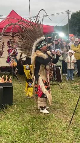 A medium shot video captures a performance outdoors in a grassy area. Two individuals dressed in elaborate Native American-inspired regalia are the main focus. They both wear large headdresses adorned with numerous feathers, intricate beadwork, and fur accents. The costumes feature layered fringed skirts, decorated sashes, and ceremonial necklaces. The person in the foreground, center, is actively playing a flute or similar wind instrument, while facing the audience. The second performer, to the left and slightly behind, also holds a flute and mirrors some of the movements. In the background, a red tent and display racks with various crafts are visible. A crowd of spectators, including men, women, and children, stands observing the performance, some of whom are holding their phones up, likely to record. The overall atmosphere appears to be that of a cultural festival or gathering, with an overcast sky suggesting a cooler climate.