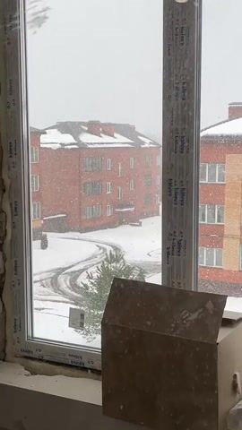 The video captures a view from inside a room, looking out of a large window onto a snowy outdoor scene. Heavy snowfall is occurring, with numerous large snowflakes visible against the grey, overcast sky.

Outside the window, two multi-story brick buildings are prominent. The building on the left is entirely red brick, while the one on the right features alternating sections of red and yellow brickwork. Both buildings have snow-covered roofs and visible chimneys. The ground between the buildings is covered in a thick blanket of snow, with tire tracks curving through it. A small evergreen tree, partially covered in snow, stands near the center of the frame, and a white car is partially visible, also blanketed in snow, to the right of the tree.

In the immediate foreground, part of a white window frame is visible. The word "kaleva" is repeatedly printed in black text on the white frame, running vertically along both the left and right sides of the window panes. On the windowsill, a cardboard box is present, and part of an aluminum can is visible to its right.

The scene depicts a winter day, likely in Подчерково, Russia, given the provided location context, with ongoing significant precipitation. No people are visible in the video.