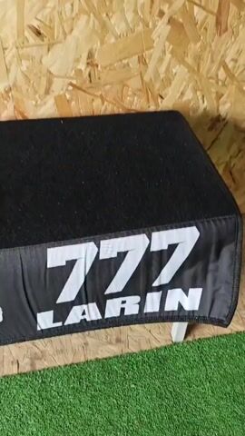 The video shows a small shed. Initially, there is a black padded step stool with a white "777 LARIN" logo on the front. The camera then pans up to show a black and white motorcycle helmet with "ORZ" printed on the side hanging on a wall made of plywood. The camera then pans across a wall displaying several motorcycle helmets in different colors and designs hanging on hooks. The shed appears to be related to "BSE LARIN" and possibly involves renting or selling motorcycle helmets.