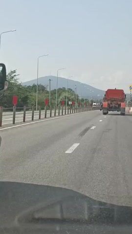 A white van drives on a highway in a sunny day. The van is in the left lane. A large orange truck with a trailer is in the right lane ahead of the van. The highway has two lanes in each direction. There are guardrails and traffic cones along the side of the road. Mountains can be seen in the background.