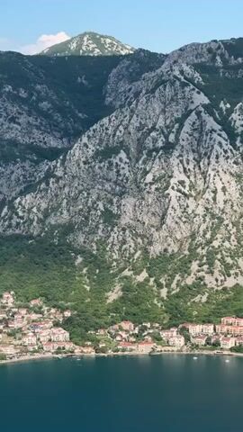 A vertical video shot from above showcases a coastal town nestled at the base of a majestic mountain range. The turquoise water of the bay stretches across the bottom of the frame, with a few sailboats visible. The town features a cluster of buildings with red-tiled roofs. The mountain behind the town is characterized by rugged, grey rock faces interspersed with green vegetation, creating a dramatic backdrop. The sky is a clear, pale blue. The video appears to be a slow pan or a static shot of this scenic landscape.