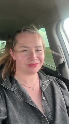 A woman with light hair, pink lipstick, and gold hoop earrings sits in the passenger seat of a moving car, wearing a dark grey denim shirt with a silver necklace. Her hair blows in the wind as she looks around and moves her mouth, possibly singing. Through the car window, a bright green field blurs past, indicating the vehicle is in motion.
