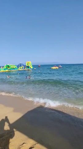 The video shows a beach scene on a sunny day. In the foreground, there is a sandy beach with lounge chairs and umbrellas. There are people relaxing on the chairs and walking on the beach. In the background, there is a body of water with a floating inflatable water park, and several people are swimming and playing in the water. The sky is clear and blue. The video pans across the beach, showing more lounge chairs and umbrellas, and then zooms in on the water park and the people enjoying the water. There are also some inflatable toys in the foreground.