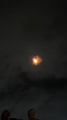The video shows a fireworks display at night. Two people are visible at the bottom of the frame, silhouetted against the dark sky. A single firework is exploding in the center of the frame, emitting a bright light and sparks. The sky is dark and hazy, with smoke from the fireworks obscuring some of the background. The firework appears to be a chrysanthemum or peony type, with a shell bursting into many smaller lights that trail down. The camera is steady, indicating it might be mounted on a tripod or held by someone standing still.