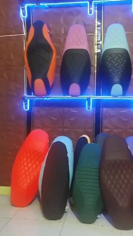 The video shows a display of motorcycle seats in a store. The seats are arranged on two shelves and on the floor. The seats are in various colors, including orange, pink, blue, purple, red, green, brown, and black. Some of the seats have a quilted pattern. The store appears to be well-lit with blue neon lights accentuating the shelves. The walls of the store have a textured brown finish.