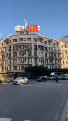 The video captures a street scene featuring a tan-colored, multi-story building with a prominent McDonald's sign on its roof. The sign is red and yellow, displaying the McDonald's logo along with a directional arrow. The building is located at an intersection, with cars and vans moving along the street. Pedestrians can be seen walking on the sidewalks. The sky is clear and blue, suggesting it is daytime. The video appears to be taken from a stationary position on the sidewalk.