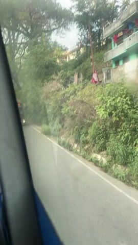 The view from inside a moving vehicle shows a bus approaching on a paved road with a solid white line on the left. The bus is bronze-colored and displays "COME TO LAHAUL" on its front display.  To the right of the road is a steep embankment covered with vegetation.  A two-story building with a light blue exterior is situated on the embankment. A person wearing a pink and white patterned outfit stands on the stairs leading to the building.  Tall trees border the scene, contributing to a dense, verdant landscape.