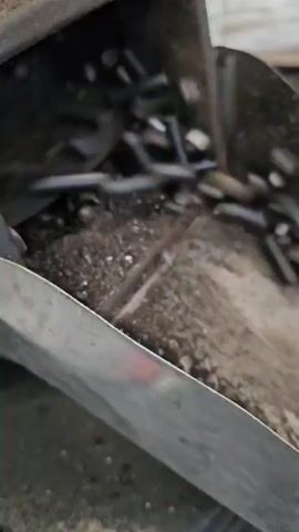 A close-up shot shows a metal trough with some debris and residue. The trough appears to be part of a larger industrial process, possibly for moving or sorting materials. The camera then pans to reveal a large bin filled with small, dark, cylindrical pellets. The bin has a perforated metal wall, allowing a view of the accumulated pellets. Towards the end of the clip, a fan or some sort of mechanical apparatus is visible in the background, suggesting ongoing operation of the machinery. There are no people or animals visible in the video.
