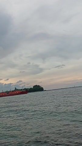 The video shows a body of water with a red ship labeled "LPG" in the distance to the left. Across the water is a small landmass with trees. The sky is overcast with grey clouds and a hint of sunset in the distance. There are small waves on the water.