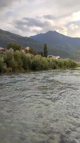 The video shows a wide, clear river flowing in front of a mountain range under a cloudy sky. The riverbed is visible with rocks and pebbles. On the riverbank, there is lush green vegetation, trees, and some buildings in the distance. The camera pans slowly from left to right, capturing the serene natural landscape.