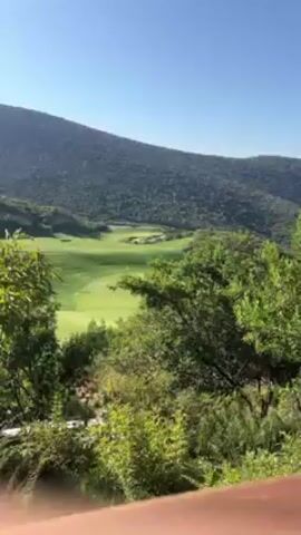 The video shows a scenic view of a golf course nestled in a valley surrounded by lush green mountains under a clear blue sky. The camera pans slightly to the right, offering a wider perspective of the course and the mountainous backdrop. In the foreground, there are trees and shrubbery, partially obscuring the view from what seems to be a balcony or deck, indicated by a wooden railing at the bottom of the frame.