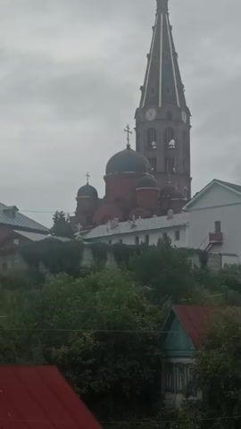 The video shows a tall, slender, red brick bell tower with a green, gold-tipped spire, featuring a clock face. To the left of the tower are several red-domed buildings topped with crosses, indicative of a church or monastery complex. A white, multi-story building with windows is situated in front of these structures. The complex is perched on a hill, partially obscured by lush green trees and foliage. In the foreground, glimpses of residential buildings with red roofs and wooden fences are visible, suggesting a village or town setting below the religious site. The sky is overcast and grey. The camera appears to be stationary, capturing a static view of the scene. There are no people visible in the frame.