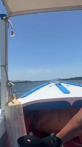 The video shows the view from the front of a small motorboat moving across a body of water. The boat has a white hull with blue trim. A white pole with a rope and a tan fender are visible on the left side of the frame. In the distance, there's a line of green trees and mountains against a clear blue sky with a few scattered clouds. The water is dark and rippled.
